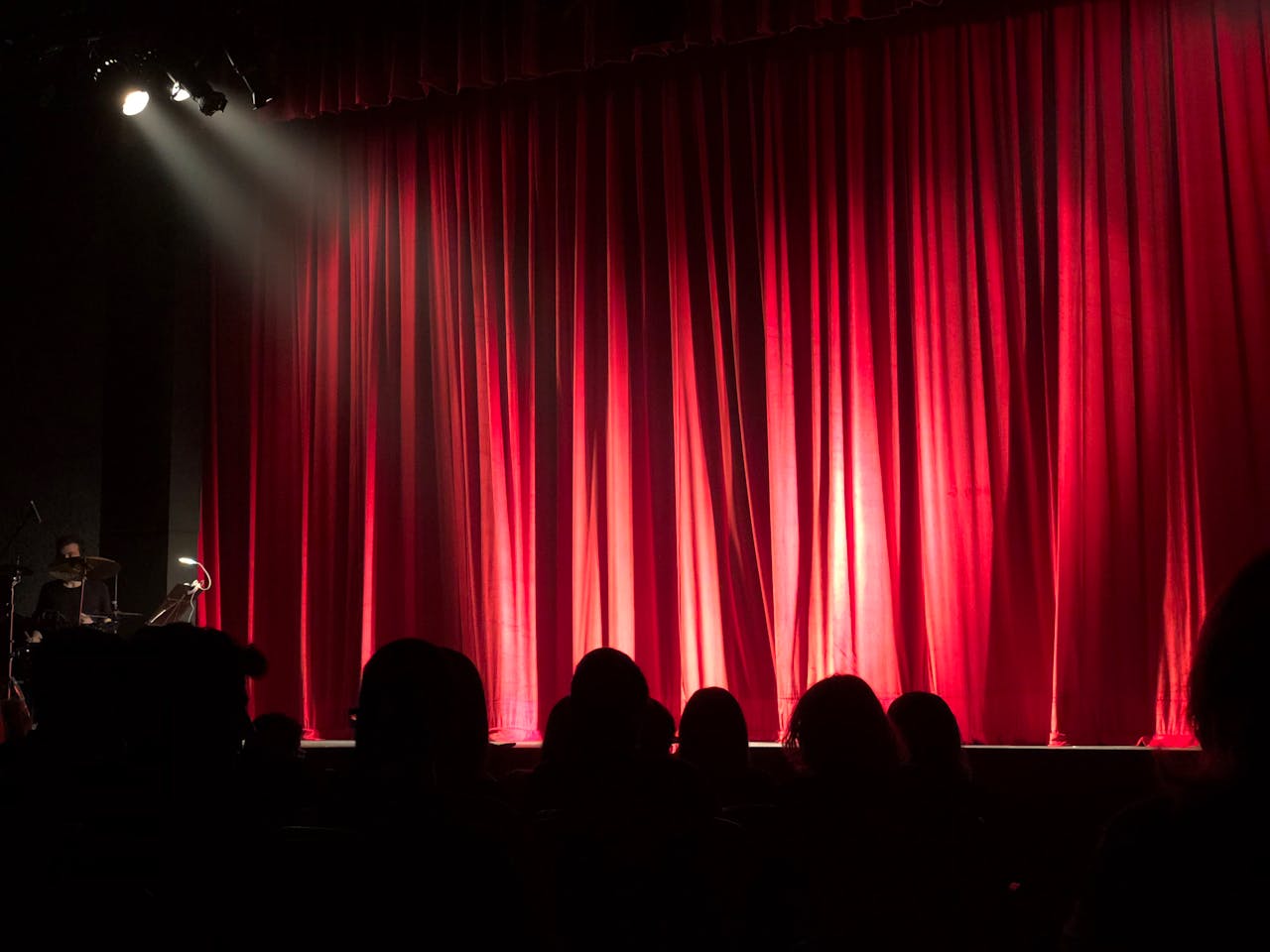 Startpagina Dimly lit theater stage with red curtains and audience silhouettes under spotlights.