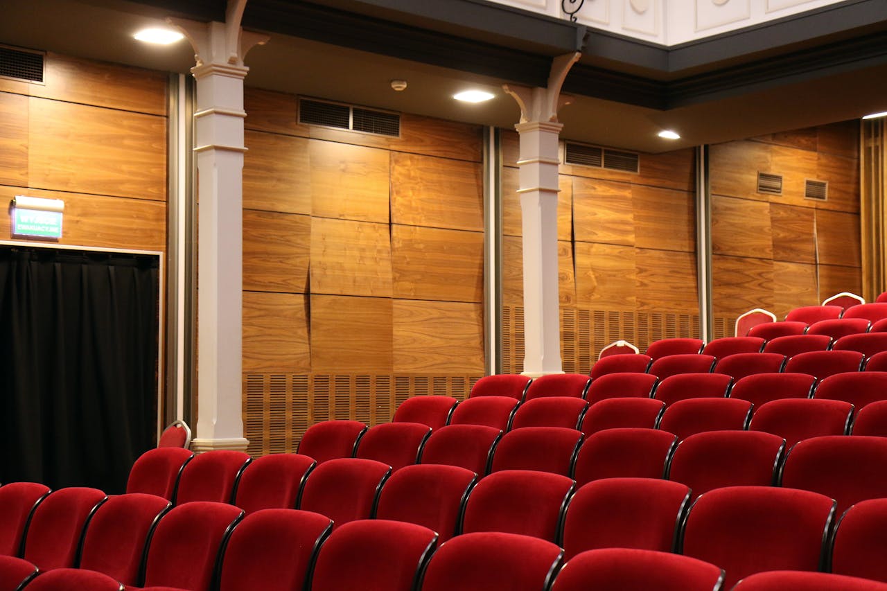 Startpagina Interior of an empty auditorium with red seats and wooden panels, lit overhead.